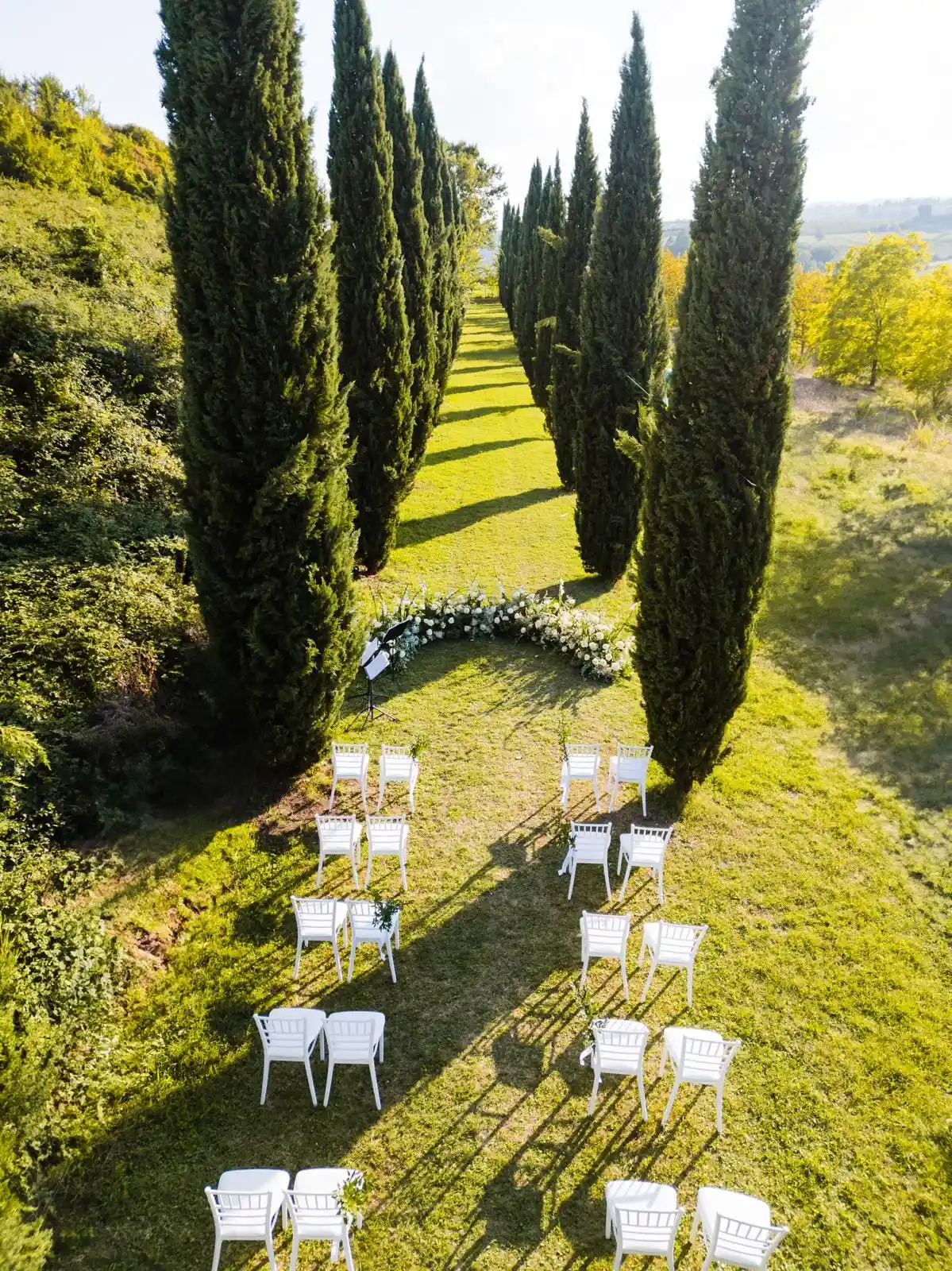 Sedia bianca elegante allineata su sentiero verde con cipressi per cerimonia all'aperto, perfetto per un Destination Wedding