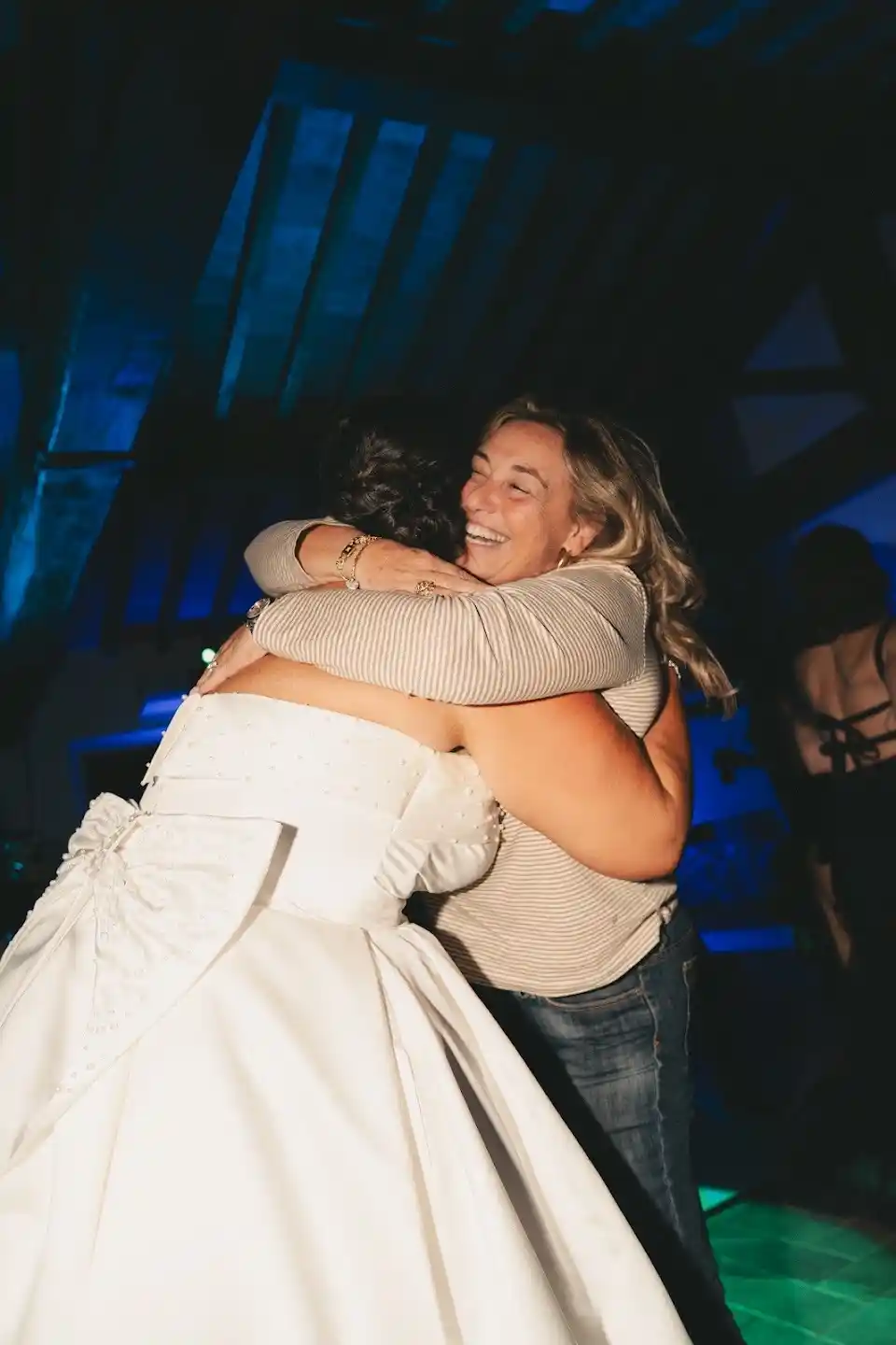 Due donne che si abbracciano sorridenti durante un evento notturno, una con abito da sposa elegante in un ambiente festoso