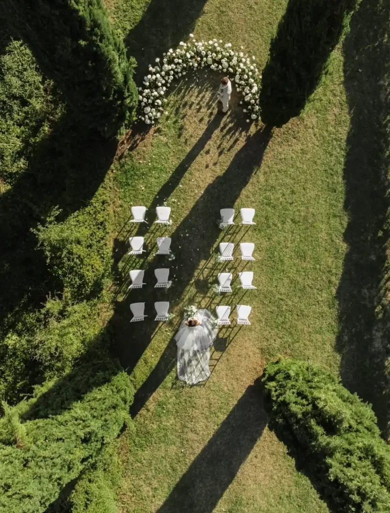 Sposi che camminano lungo un sentiero verde con archi di fiori bianchi e sedie per ospiti in un contesto naturale per Eventi per matrimoni