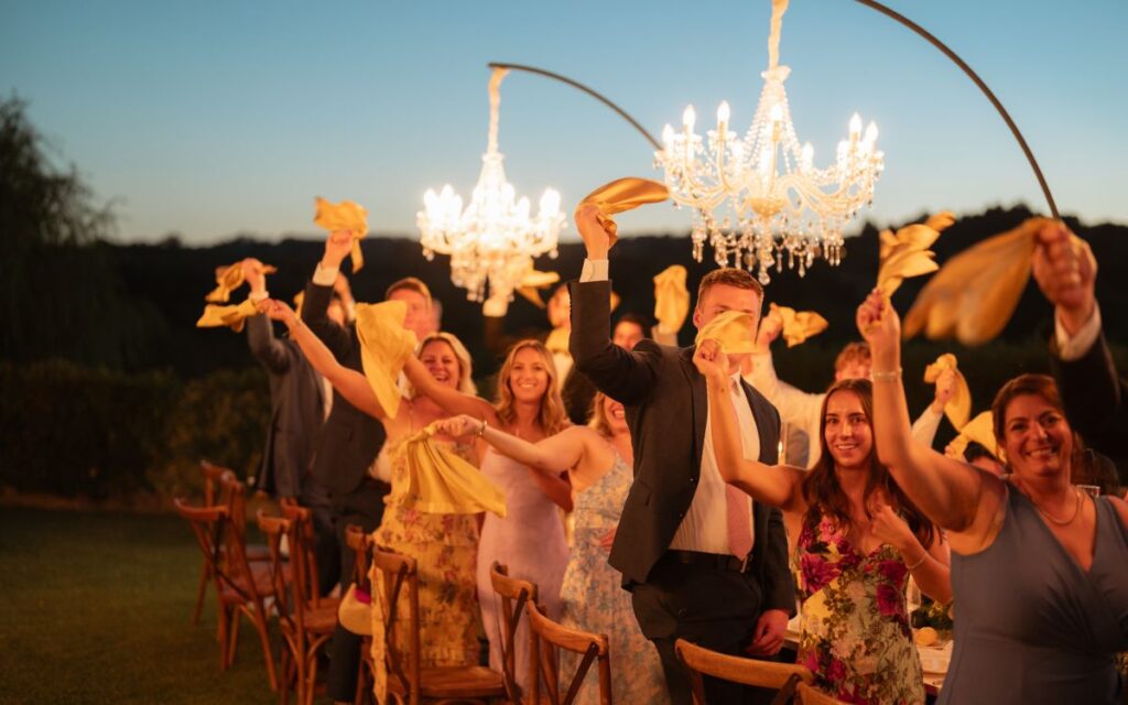 Gruppo di sposi e invitati che alzano lanterne luminose durante un evento serale all'aperto, con sfondo di candelabri e natura