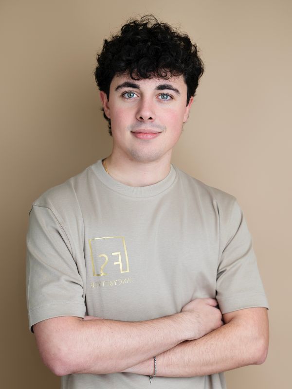 Ragazzo sorridente con maglietta beige e logo dorato, posa sicura con braccia incrociate