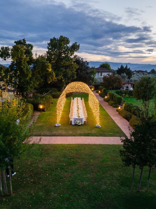 Cerimonia matrimoniale esclusiva in giardino toscano con archi di luci e tavoli all'aperto al tramonto