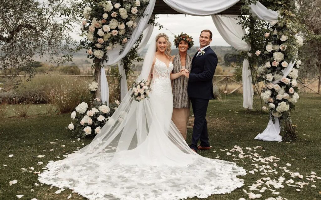 Sposa con abito da matrimonio bianco sotto un arco floreale all'aperto, accompagnata da testimoni e sposo in giacca scura, location per matrimoni romantica
