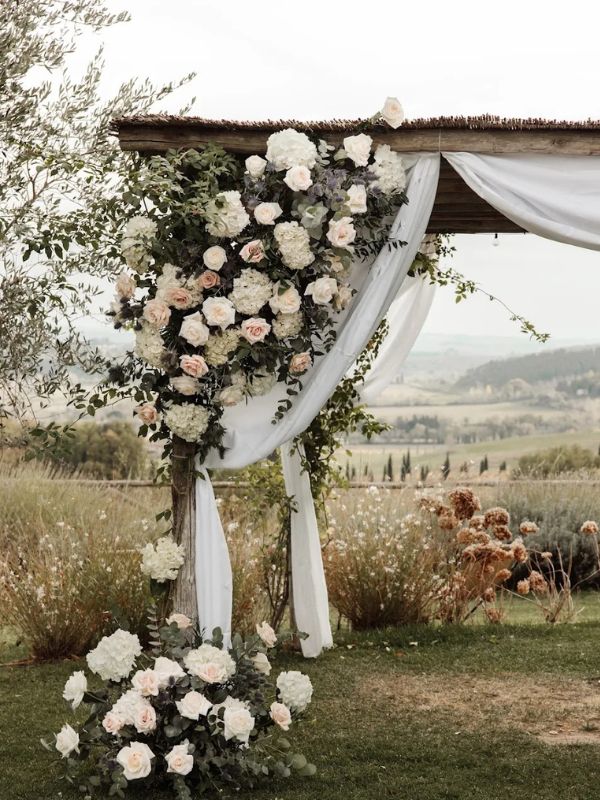 Arco floreale romantico per cerimonia all'aperto con rose bianche e verdi, sfondo paesaggio collinare