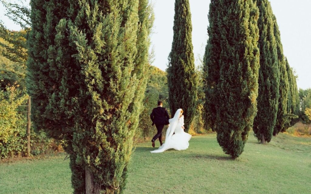Sposi che camminano tra alberi di cipresso in un giardino verde per cerimonia nuziale all'aperto, ideale per un evento esclusivo organizzato da un'Agenzia Wedding Planner