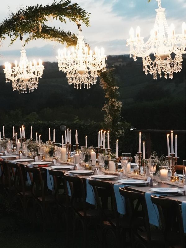 Tavoli eleganti con tovaglie blu e candelabri luminosi per un evento serale all'aperto, decorazioni floreali e lampadari cristallo